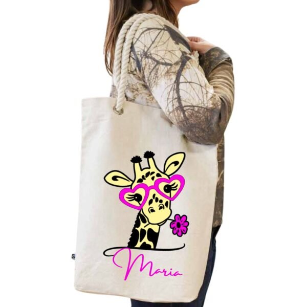 Tote bag blanco de algodón con asa de cuerda blanca y personalizado con la imagen "cuqui" de una jirafa con gafas y una flor de color fucsia conjuntamente con el nombre que se quiera personalizar tambien en el mismo color fucsia.